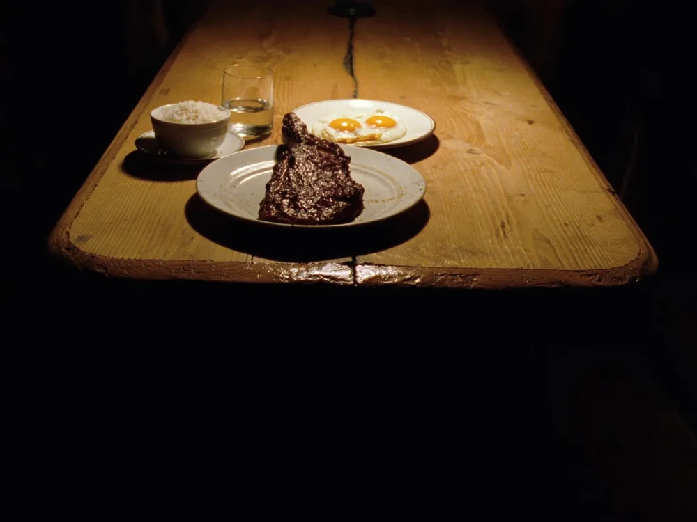 Uma mesa de madeira com carne, ovos, arroz e um copo d'água sob uma luz quente